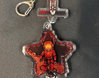 May include: A clear acrylic keychain featuring a red astronaut in a spacesuit on a red star-shaped background. The keychain has a silver keyring and a cross-shaped charm with a rocket design.