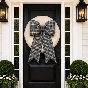 May include: A round, cream-colored textured wreath featuring a large, black and white plaid fabric bow with long, pointed tails. The wreath is centered on a black front door, flanked by two black lanterns and two black planters with green shrubs.