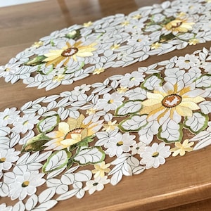 May include: A set of four white embroidered table runners with yellow sunflower designs. The runners are placed on a wooden table with a green plant in a pot in the center.