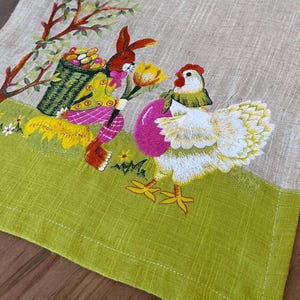 May include: A beige linen table runner with green borders features embroidered images of a bunny carrying a basket of eggs and a chicken holding a pink egg. The bunny is wearing a pink and green outfit and the chicken is white with a red comb.