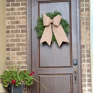 May include: A dark brown wooden door features a green evergreen wreath adorned with a large, tan and white gingham check fabric bow. To the left, a black planter holds green foliage with red flowers, sitting atop a gold-toned metal lantern.