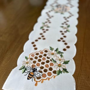 May include: White embroidered table runner with a honeycomb design, a bee, and white flowers. The runner has a scalloped edge.