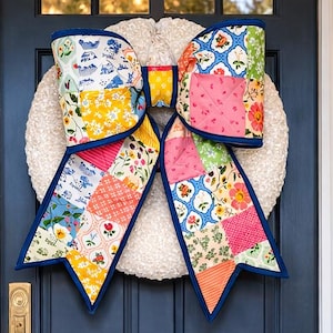 May include: A navy blue front door with a large, colorful bow-shaped wreath. The wreath is made of a white, fluffy material and the bow features a patchwork design with various floral and geometric patterns. Potted plants with pink and blue flowers flank the door.