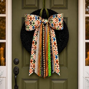 May include: A black textured wreath hangs on an olive green door, featuring a large bow with a diamond pattern of colorful Halloween icons. Long, multi-textured ribbons in orange, green, black, and gold dangle from the center of the bow.
