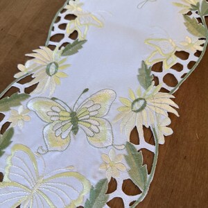 May include: White table runner with embroidered green leaves and yellow butterflies. The runner has a scalloped edge and a delicate lace pattern.