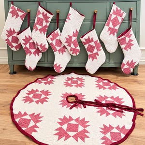 May include: A set of seven quilted Christmas stockings and a matching circular tree skirt. Each item features a white background with a repeating dark pink geometric star pattern, finished with dark pink fabric trim and hanging loops.