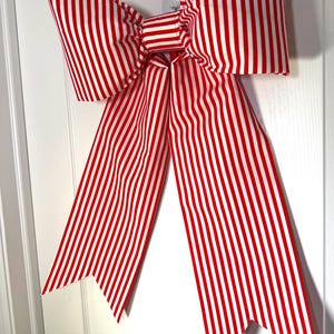 Puede incluir: Un gran lazo decorativo con rayas verticales rojas y blancas. El lazo es de tela y tiene largas colas puntiagudas. Está fijado a una puerta blanca, creando un acento festivo y llamativo.