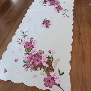 May include: White table runner with pink embroidered flowers and a small bird. The runner has a scalloped edge.