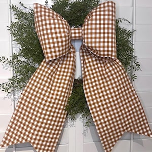 May include: A large brown and white gingham bow with a center loop. The bow is tied around a wreath of green foliage.