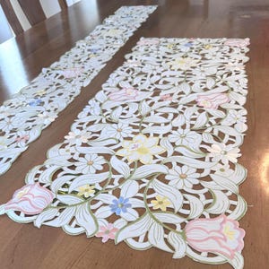 May include: Two white embroidered table runners with a floral design. The runners feature a delicate lace pattern with pink, yellow, and blue flowers.