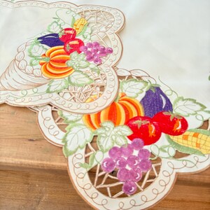 May include: Cream-colored embroidered table runner with a harvest theme. Features colorful embroidered pumpkins, tomatoes, grapes, corn, and eggplant. The edges are scalloped with a decorative brown border.