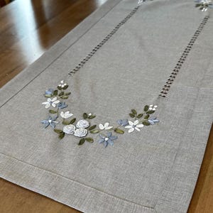 May include: A gray linen table runner with a delicate floral embroidery design featuring white and blue flowers and green leaves. The runner has a stitched border.