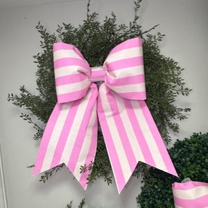 May include: A large pink and white striped bow with a pointed end on each side. The bow is made of fabric and is tied around a green wreath.