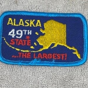 May include: A rectangular patch with a blue border and background. The patch features the word "ALASKA" in yellow, "49TH STATE" in white and red, and a yellow outline of Alaska. The text "...THE LARGEST!" is in red.