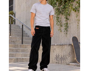 Puede incluir: Una camiseta de manga corta y cuello redondo en color gris claro combinada con pantalones de pana negros de corte holgado con una pequeña etiqueta rectangular naranja en el bolsillo delantero, junto a zapatillas bajas blancas y negras con cordones.