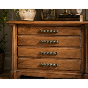 Peut inclure: Une commode en bois dotée de quatre tiroirs horizontaux avec une texture de grain de bois visible. Chaque tiroir est équipé d'une longue poignée métallique en forme de fuseau, avec une finition sombre et patinée et un motif segmenté.