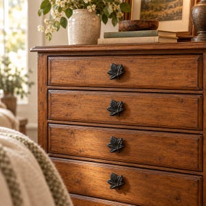 May include: A wooden dresser with four drawers, each featuring a dark metal leaf-shaped pull. The dresser has a warm brown finish with visible wood grain patterns. The drawer fronts are framed with a simple rectangular molding detail.