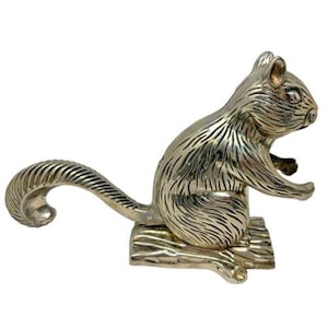 May include: A silver-colored squirrel figurine, detailed with textured fur and a curled tail. The squirrel is posed sitting on a log-shaped base. The figurine is made of metal and has a shiny finish.