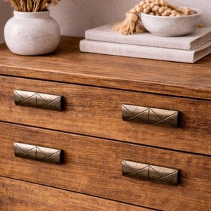 May include: Close-up of a wooden dresser with four drawers. Each drawer has a bronze-colored, geometric handle. The wood grain is visible, and the finish is a warm brown. A vase and books are visible on top.