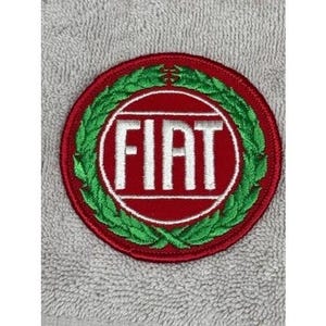 May include: Embroidered patch featuring the Fiat logo. The patch is circular, with a red background and a white Fiat logo in the center. A green laurel wreath surrounds the logo. The patch is likely for clothing or accessories.