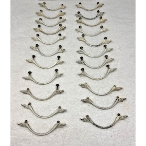 May include: A collection of vintage silver-toned drawer pulls. Each pull has a curved handle with decorative leaf-like accents on the ends and a small, dark knob in the center. The pulls are arranged in neat rows.