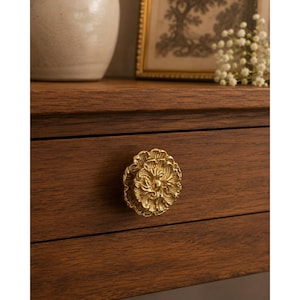 May include: Close-up of a gold-colored, floral-shaped drawer pull on a wooden cabinet. The drawer pull has detailed petal designs. The cabinet is made of dark brown wood. A vase and framed picture are in the background.