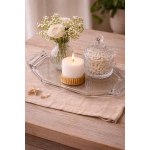 May include: A decorative arrangement featuring a lit white candle on a gold base, a small glass vase with white flowers, and a glass jar with a lid. The items are displayed on a clear, geometric tray and a beige linen runner.
