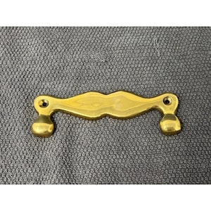 May include: A brass-colored cabinet pull with a curved center and rounded ends. The hardware has two mounting holes and is set against a textured gray surface. This decorative pull is suitable for furniture restoration or new builds.
