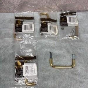 Pode incluir: Várias puxadores e puxadores de armário de latão novos em embalagens de plástico transparente são exibidos em uma superfície cinza claro. O hardware inclui várias formas e tamanhos, com parafusos visíveis em alguns. A estética geral é moderna e funcional.