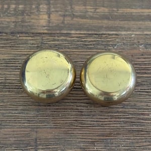 Può includere: Due pomelli circolari per armadietti in ottone lucido con una superficie liscia e riflettente. Ogni pomello presenta una parte superiore arrotondata a cupola e una finitura color oro metallizzato con lievi graffi superficiali.