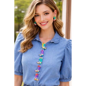 May include: A denim shirt with colorful cowboy boot-shaped buttons and matching earrings. The buttons are in various colors, including yellow, purple, and turquoise, with black and white cowhide patterns. The earrings have a similar design.
