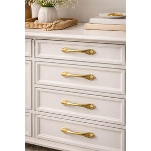 May include: A white chest of drawers with gold-coloured drawer pulls. The pulls have an ornate design. The chest is in a room with a vase of flowers and other decorative items on top.