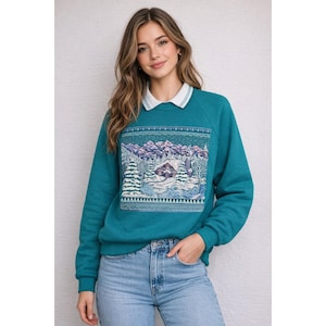 May include: Teal sweatshirt with a white collar and a winter landscape design. The design features a cabin, trees, and mountains in shades of blue, white, and green. The sweatshirt is paired with blue jeans.