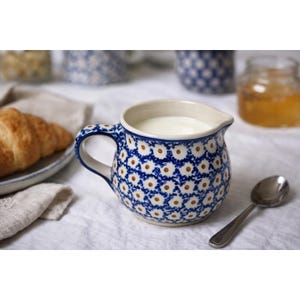 May include: A blue and white patterned ceramic milk jug, filled with milk. The jug has a handle and a spout. A croissant, a spoon, and a jar of honey are also visible.