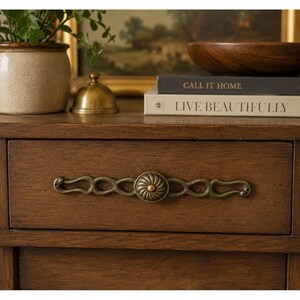 May include: A wooden drawer featuring an ornate, elongated brass handle with a central circular rosette design. The drawer is part of a brown wooden piece of furniture, positioned below two stacked books titled CALL IT HOME and LIVE BEAUTIFULLY.