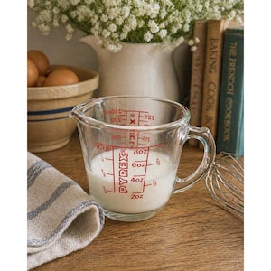 Puede incluir: Un vaso medidor Pyrex transparente con marcas rojas, que contiene un líquido blanco. El vaso tiene un asa y está parcialmente lleno. Un paño a rayas y un batidor de varillas están en primer plano. Un cuenco con huevos y libros de cocina están en el fondo.