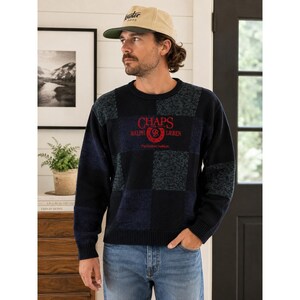 Könnte beinhalten: Ein langärmeliger Pullover mit Rundhalsausschnitt und einem dunkelblauen und schwarzen Karomuster. Auf der Brust befindet sich der rote Stickschriftzug CHAPS RALPH LAUREN mit einem kreisförmigen Logo und dem Text The Authentic Tradition.