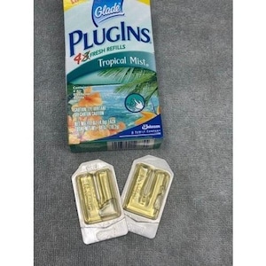 Può includere: Una confezione di ricariche per deodorante per ambienti Glade PlugIns Tropical Mist con due ricariche singole. La confezione presenta una scena tropicale con palme e fiori. Le ricariche sono in un liquido trasparente e giallo.