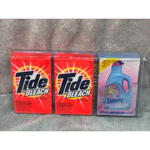 May include: A selection of sample-sized laundry products in a clear wrapper. Two red boxes of Tide with Bleach laundry detergent and a blue and pink box of Downy Ultra fabric softener. The Tide boxes have a white and yellow burst design.