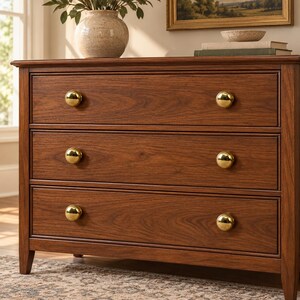 May include: A wooden dresser with a rich brown finish featuring three horizontal drawers. Each drawer is equipped with two polished gold-toned spherical knobs. The piece has a rectangular frame with clean lines and a smooth, visible wood grain texture.