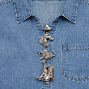 May include: Close-up of a denim shirt with silver-colored cowboy-themed buttons. The buttons include a cowboy hat, a horse head, a saddle, and a pair of cowboy boots. The shirt has a chest pocket on the right side.