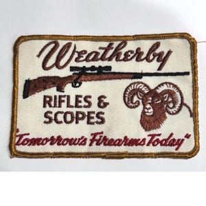 Può includere: Toppa ricamata con la scritta "Weatherby" sopra un fucile e le parole "RIFLES & SCOPES". Una testa di ariete è a destra. Il testo in basso recita "Tomorrow's Firearms Today". La toppa ha un bordo dorato.