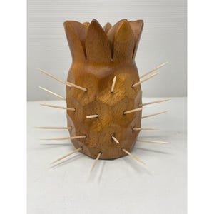 Könnte beinhalten: Ein Zahnstocherhalter aus Holz in Ananasform mit natürlicher Oberfläche. Der Halter ist mit einer strukturierten Oberfläche geschnitzt und weist mehrere Zahnstocher auf, die an den Seiten eingesetzt sind und an die stachelige Außenseite der Frucht erinnern. Die Oberseite ist wie eine Ananaskrone geformt.
