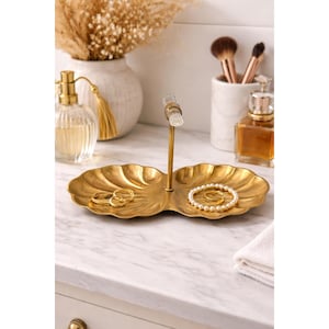 May include: A gold-colored, two-sectioned decorative tray in a shell design, holding rings and a pearl bracelet. A clear, vertical jewelry holder is in the center. The tray sits on a marble surface, with perfume bottles and brushes in the background.