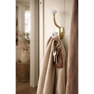 May include: A brass wall hook with white ceramic knobs holds a beige scarf and a set of keys with a brown leather fob. The hook is mounted on a cream-colored door, with a glimpse of a room in the background.