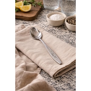 May include: A stainless steel spoon with a decorative handle rests on a beige linen napkin. The spoon has an engraved floral design. The background includes small bowls of salt and pepper, and a wooden cutting board with lemon slices.
