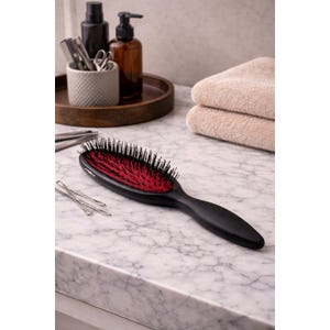 May include: A black hairbrush with a red cushion and black bristles. The brush is on a marble countertop, with bobby pins, towels, and toiletries in the background. The brush is approximately 9 inches long.