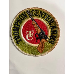 Puede incluir: Parche bordado con el texto "THOMPSON CENTER ARMS" en un diseño circular. El parche presenta una ilustración de rifle, un círculo rojo y un borde verde. El parche tiene un fondo color crema.