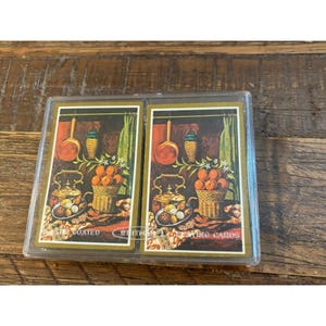 May include: Two decks of Whitman playing cards in a clear plastic case. The card backs feature a still life illustration of oranges in a woven basket, a copper kettle, and a hanging copper pan against a dark background with green curtains.