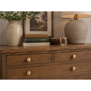 Puede incluir: Una cómoda de madera con cuatro cajones y pomos dorados pulidos. Encima hay un jarrón beige texturizado con vegetación, una lámina de paisaje enmarcada, tres libros apilados titulados The Pattern of Industry y Annie Leibovitz At Work, y una lámpara.